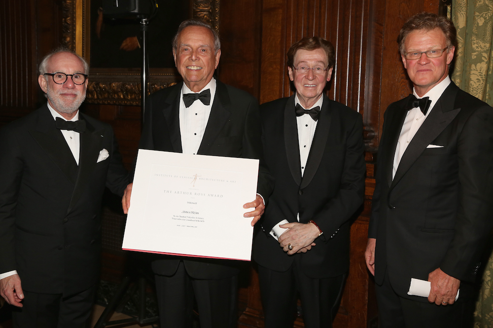 Four people wearing black tuxedos facing forward. One of them is holding an award certificate.