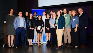 A group of people on a stage gathered together, smiling, accepting an award.