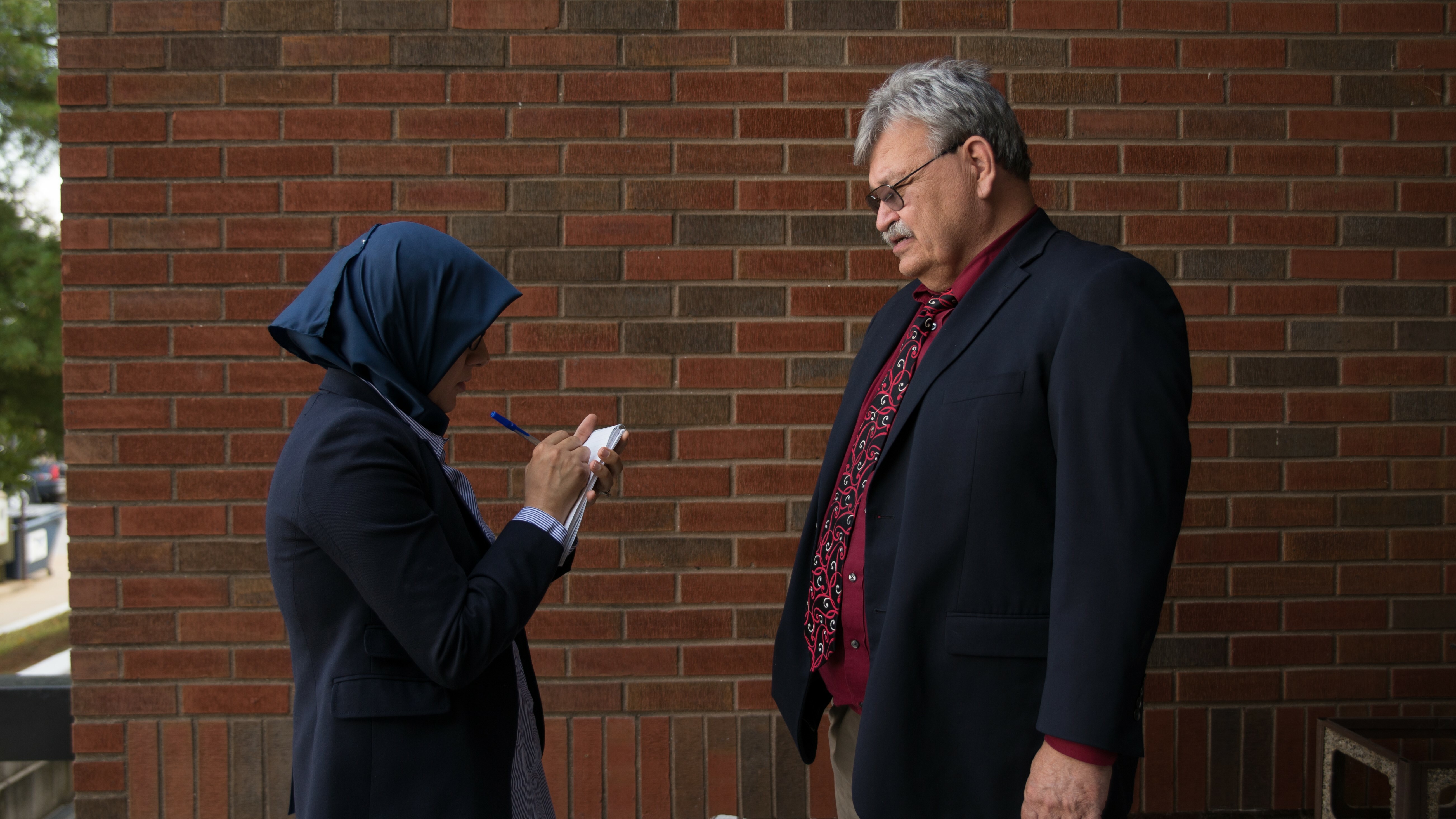 Two people facing each other. One speaking and the other is a reporter taking notes.