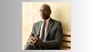 A person sitting on a bench wearing a suit and eyeglasses, smiling at the camera.