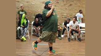 A person wearing a green shirt and camouflage shorts holding a microphone walking in front of a group of people seated in white chairs.