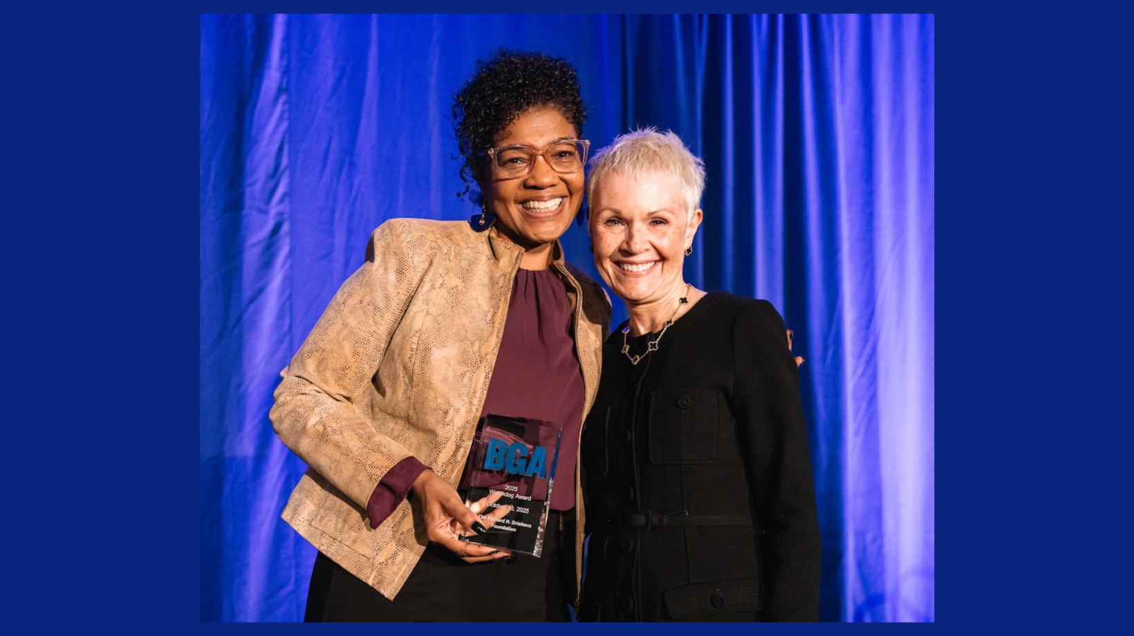 Two people facing the camera, smiling. One person is holding a glass award. The background is blue.