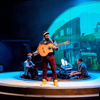 A group of musicians performing on a circular stage in front of a projection of buildings. The musician at center stage is holding a guitar.