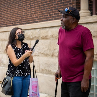 A reporter standing a holding a microphone to another standing person who they are interviewing.