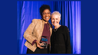 Two people facing the camera, smiling. One person is holding a glass award. The background is blue.